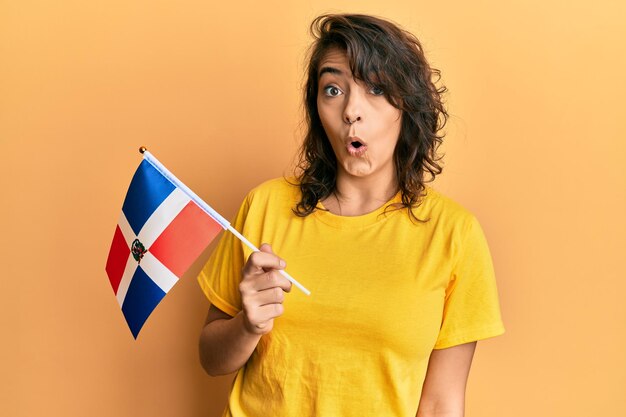 Jeune femme hispanique tenant le drapeau de la république dominicaine effrayée et étonnée avec la bouche ouverte pour la surprise, le visage d'incrédulité