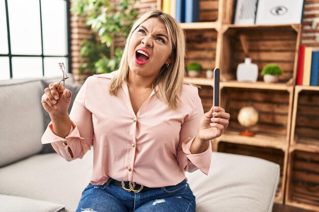 Jeune femme hispanique tenant un recourbe-cils et une lime à ongles en colère et folle criant frustré et furieux, criant de colère en levant les yeux.