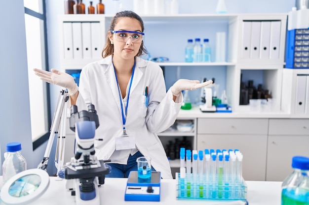 Jeune femme hispanique travaillant au laboratoire scientifique expression désemparée et confuse avec les bras et les mains levés. notion de doute.