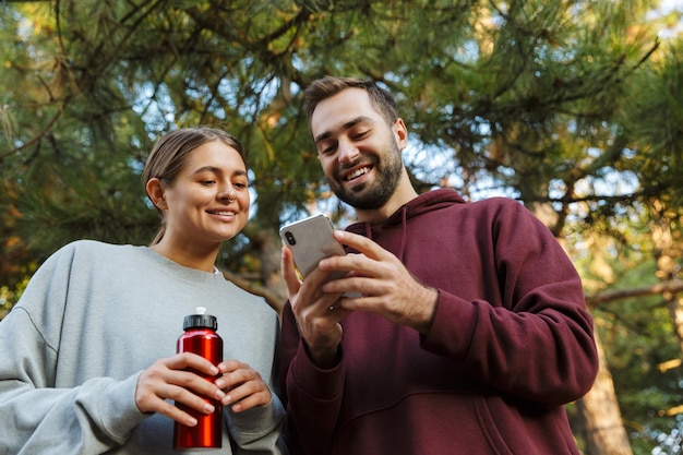 Une jeune femme et un homme sportifs positifs heureux utilisant un téléphone portable à l'extérieur dans un parc verdoyant tenant une bouteille d'eau.