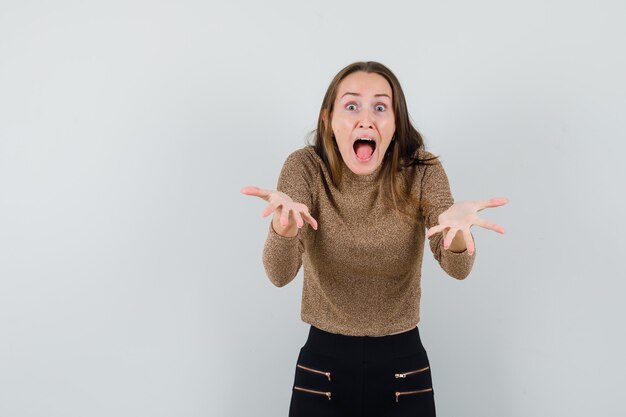 Jeune femme hurlant tout en étirant les mains vers la caméra en pull doré et pantalon noir et à la vue de face, inquiète.