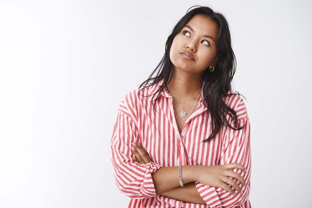 Jeune femme impatiente en détresse et sous pression regardant le coin supérieur gauche en vérifiant l'heure, les bras croisés sur la poitrine et souriant insatisfait de la file d'attente lente, posant sur fond blanc