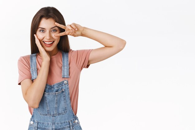 Jeune femme impertinente et joyeuse en salopette, t-shirt, montrant un signe de paix ou de victoire sur l'œil en mouvement disco, touchant doucement la joue, l'air déterminé et confiant, debout sur fond blanc, sûr de lui