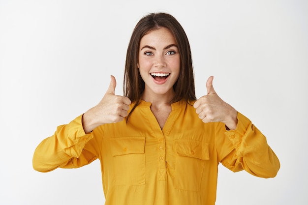 Jeune femme impressionnée montrant les pouces vers le haut et souriante étonnée, louant quelque chose de cool, debout sur un mur blanc