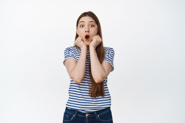 Une jeune femme impressionnée regarde avec émerveillement la mâchoire baissée, regarde excitée, se tient la main près du visage avec une émotion surprise sur blanc.