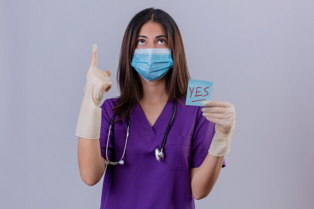 Jeune femme infirmière portant des gants de masque de protection uniforme médical et avec stéthoscope tenant un papier de rappel avec oui mot pointant avec le doigt vers le haut