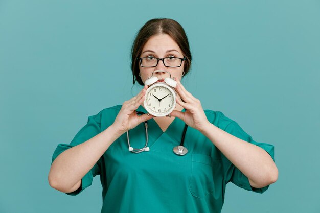 Jeune femme infirmière en uniforme médical avec stéthoscope autour du cou montrant un réveil à l'air inquiet debout sur fond bleu