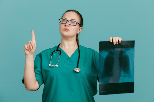 Jeune femme infirmière en uniforme médical avec stéthoscope autour du cou tenant une radiographie pulmonaire à la confiance montrant l'index ayant de bonnes nouvelles debout sur fond bleu