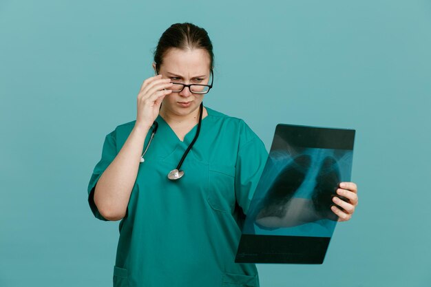 Jeune femme infirmière en uniforme médical avec stéthoscope autour du cou tenant une radiographie pulmonaire en le regardant avec un visage sérieux debout sur fond bleu