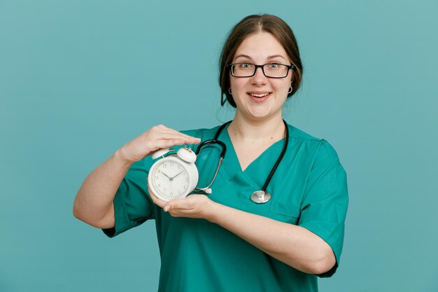 Jeune femme infirmière en uniforme médical avec stéthoscope autour du cou tenant un réveil regardant la caméra souriant confiant heureux et positif debout sur fond bleu