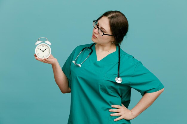 Jeune femme infirmière en uniforme médical avec stéthoscope autour du cou tenant un réveil en le regardant avec un visage sérieux debout sur fond bleu