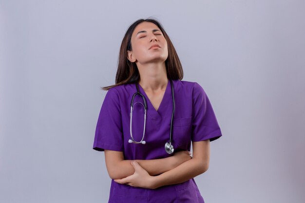 Jeune femme infirmière en uniforme médical et avec stéthoscope à l'estomac mal en point ayant des douleurs debout