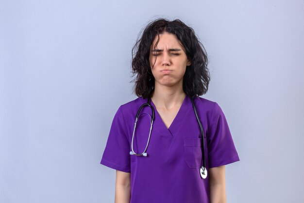 Jeune femme infirmière en uniforme médical et avec stéthoscope à la fatigue soufflant ses joues debout avec les yeux fermés sensation de fatigue