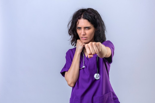 Jeune femme infirmière en uniforme médical et avec stéthoscope prêt à se battre avec le geste de défense poing visage en colère et bouleversé