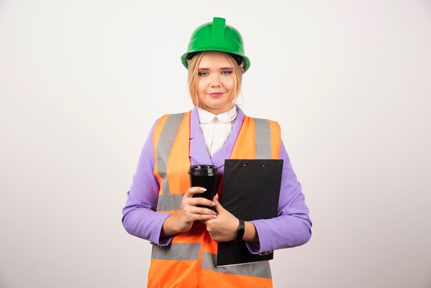 Jeune femme ingénieur industriel en uniforme avec presse-papiers et tasse noire sur blanc.