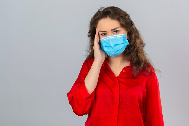 Jeune femme inquiète portant un chemisier rouge dans un masque de protection médicale à la recherche de mal avec la main près de la tête ayant des maux de tête sur fond blanc isolé