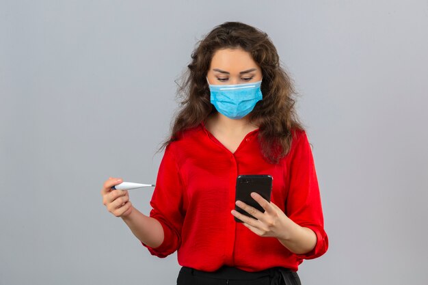 Jeune femme inquiète portant un chemisier rouge dans un masque de protection médicale tenant un thermomètre et regardant sur l'écran de son téléphone mobile sur fond blanc isolé