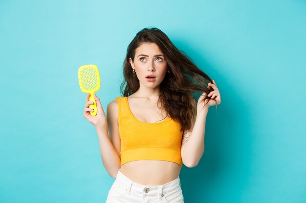 Jeune femme inquiète pour l'état de ses cheveux, montrant une brosse et regardant le coin supérieur gauche avec un visage concerné, debout sur fond bleu.