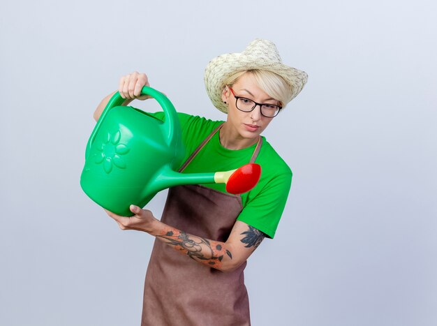 Jeune femme jardinier aux cheveux courts en tablier et chapeau tenant un arrosoir souriant confiant