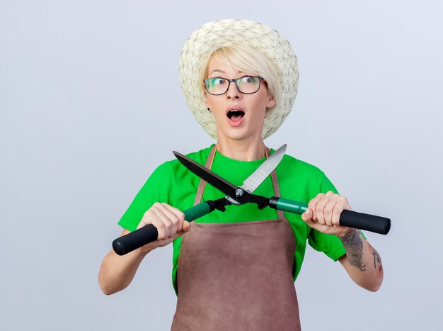 Jeune femme jardinier aux cheveux courts en tablier et chapeau tenant des coupe-haies lookign de côté d'être surpris