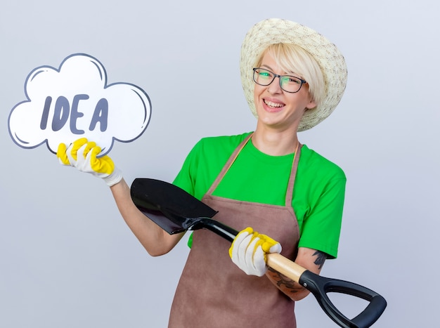 Jeune femme jardinier aux cheveux courts en tablier et chapeau tenant une pelle montrant un signe de bulle de parole avec une idée de mot souriant avec un visage heureux