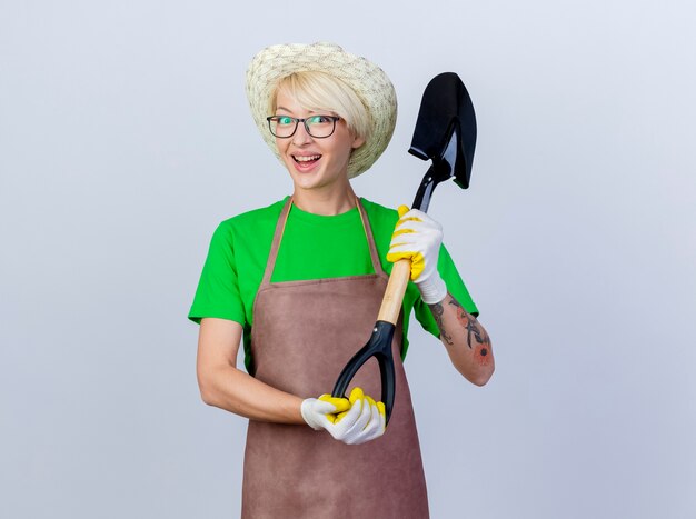 Jeune femme jardinier aux cheveux courts en tablier et chapeau tenant une pelle souriant gaiement