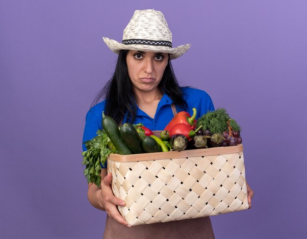 Jeune femme de jardinier confuse en uniforme et chapeau tenant un panier de légumes regardant à l'avant isolé sur un mur violet avec espace pour copie