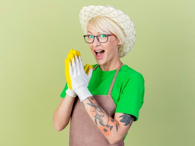 Jeune femme jardinière aux cheveux courts en tablier et chapeau portant des gants en caoutchouc tenant les paumes ensemble souriant joyeusement debout sur fond clair