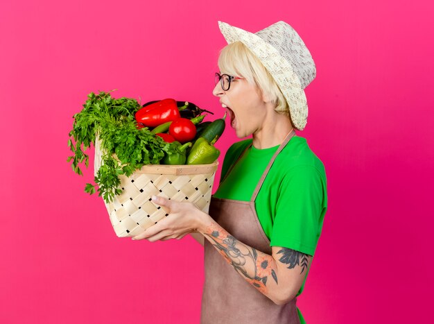 Jeune femme jardinière aux cheveux courts en tablier et chapeau tenant une caisse pleine de légumes