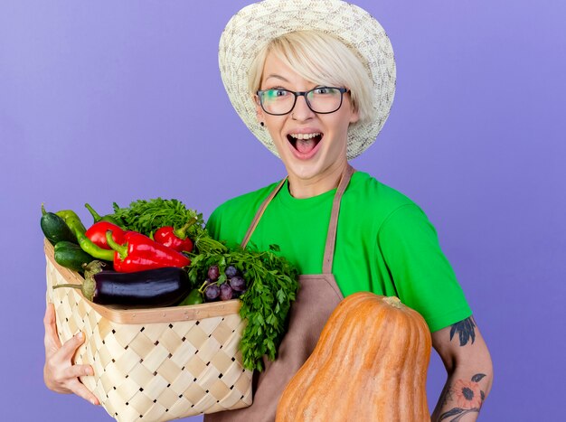 Jeune femme jardinière aux cheveux courts en tablier et chapeau tenant une caisse pleine de légumes