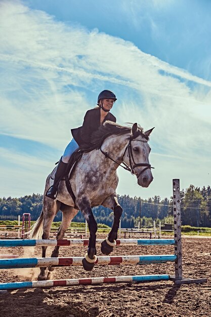 Jeune femme jockey sur cheval gris pommelé sautant par-dessus l'obstacle dans l'arène ouverte.