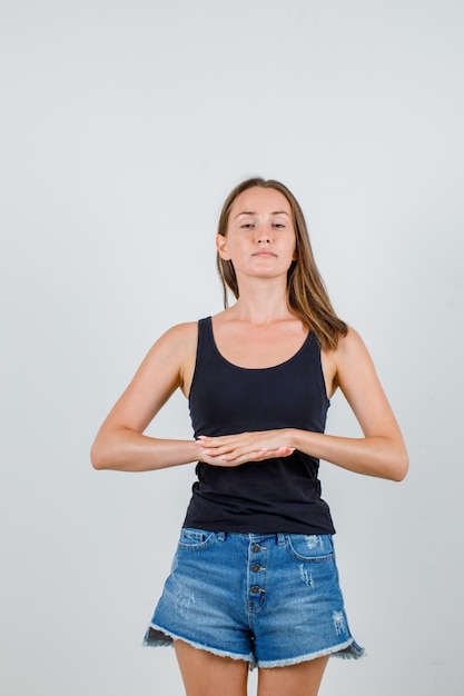 Jeune femme joignant deux mains en maillot noir, short et à la détente. vue de face.