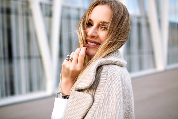 Jeune femme jolie blonde élégante posant dans la rue, look sensuel naturel, souriant et regardant la caméra, portant un manteau beige à la mode et des accessoires de luxe, printemps automne, couleurs douces.