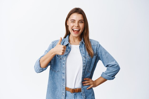 Jeune femme joyeuse aux cheveux blonds et aux vêtements décontractés, montrant le pouce en l'air, comme une bonne chose, l'approuve et le recommande, loue un bon travail ou dit oui, mur blanc