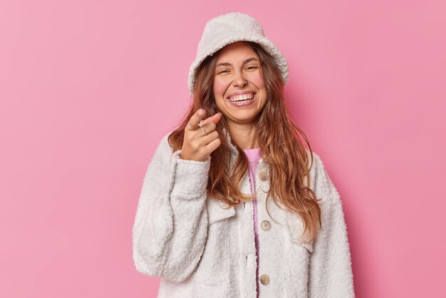 Une jeune femme joyeuse, de longs cheveux longs et ondulés, pointe l'index vers vous, remarque quelque chose de drôle devant les sourires, porte avec plaisir un manteau de fourrure et un panama isolé sur fond de studio rose. Hé rejoins-moi