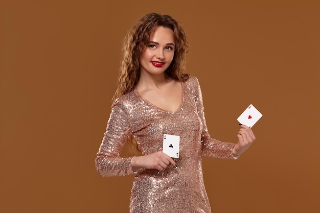 Une jeune femme joyeuse vêtue d'une robe à paillettes dorées tient deux cartes à jouer dans ses mains, souriant et regardant la caméra. Concept de casino sur fond marron.