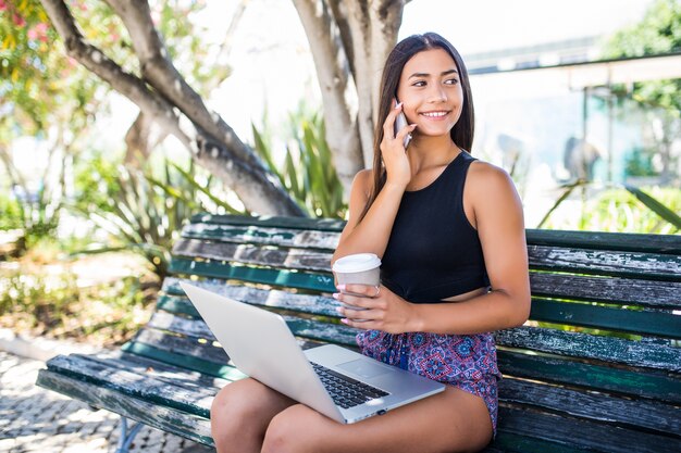Jeune femme latine assise sur un banc, parler sur smartphone, travaillant sur un ordinateur portable à l'extérieur.