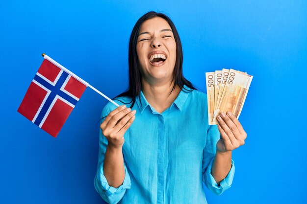 Photo gratuite jeune femme latine tenant le drapeau de la norvège et les billets en couronne souriant et riant fort à haute voix parce que drôle de blague folle