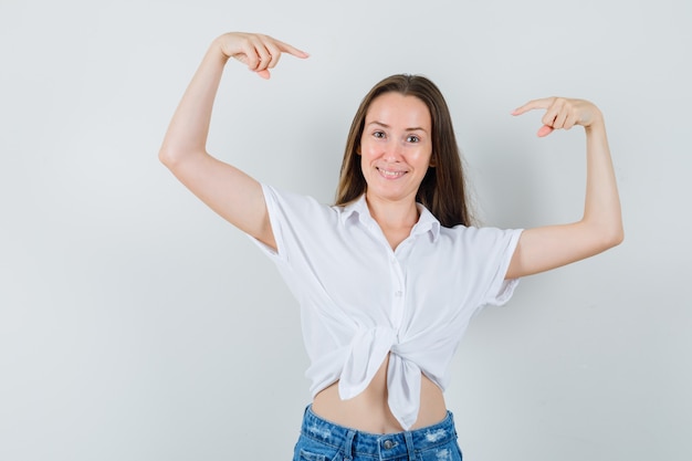 Jeune femme levant les bras tout en se pointant en chemisier blanc et à l'air heureux. vue de face.