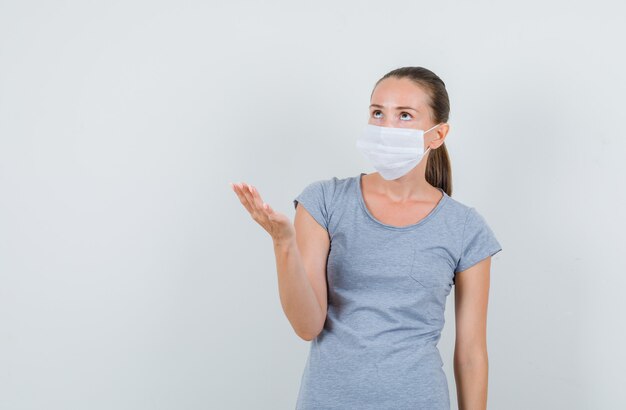 Jeune femme levant la main en geste de questionnement en t-shirt gris, masque, vue de face.