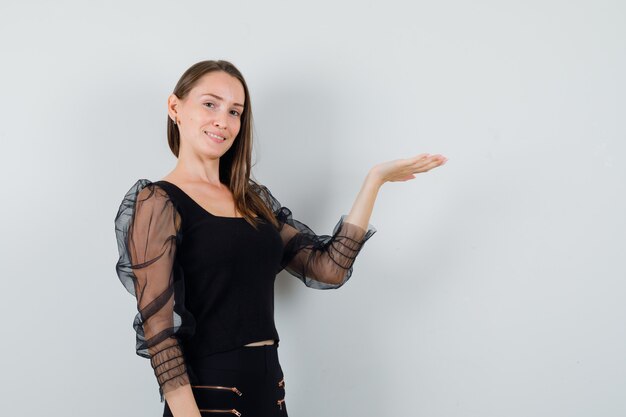 Jeune femme levant la main avec la paume ouverte de côté montrant quelque chose en chemisier noir et à la satisfaction