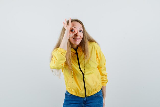 Jeune femme levant la main sur la tête en blouson aviateur jaune et jean bleu et à la pensif. vue de face.