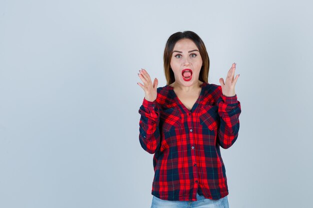 Jeune femme levant les mains en chemise à carreaux et l'air choquée. vue de face.