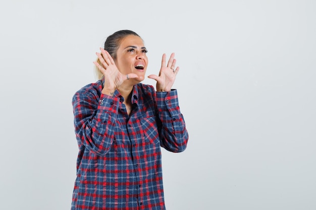 Jeune femme levant les mains comme appeler quelqu'un en chemise à carreaux et à la jolie. vue de face.