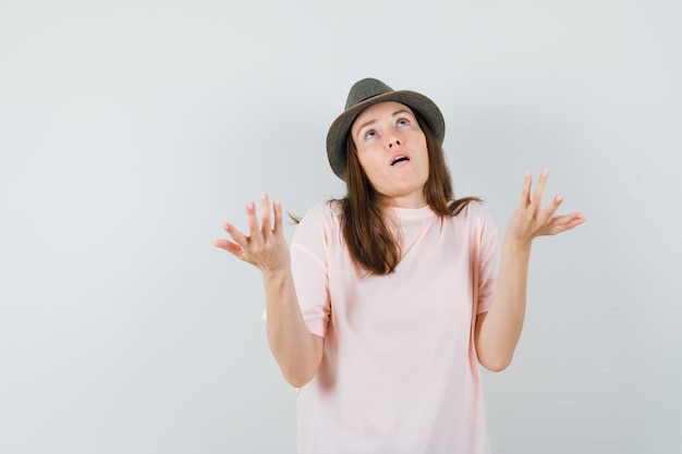 Jeune femme levant les mains de manière interrogative en t-shirt rose, vue de face de chapeau.