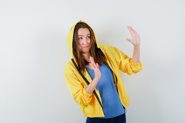 Jeune femme levant les mains de manière protectrice en t-shirt, veste et ayant l'air effrayée, vue de face.