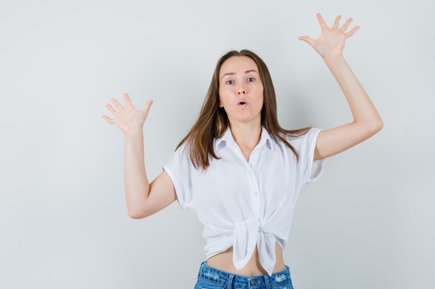 Jeune femme levant les mains avec les paumes ouvertes en chemisier blanc et regardant bizarre, vue de face.