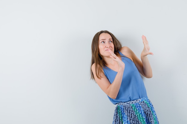 Jeune femme levant les mains pour se défendre de quelque chose en chemisier bleu, jupe et regardant anxieux, vue de face.