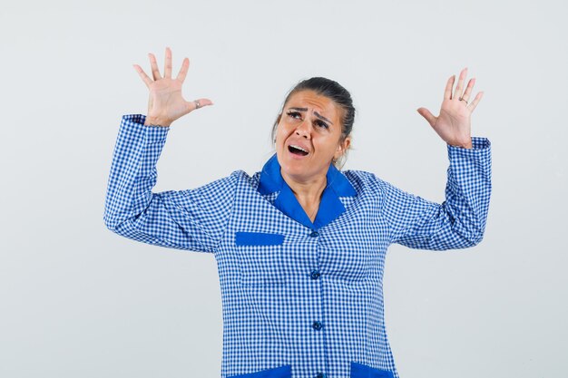 Jeune femme levant les mains en reddition pose en chemise de pyjama vichy bleu et à épuisé. vue de face.