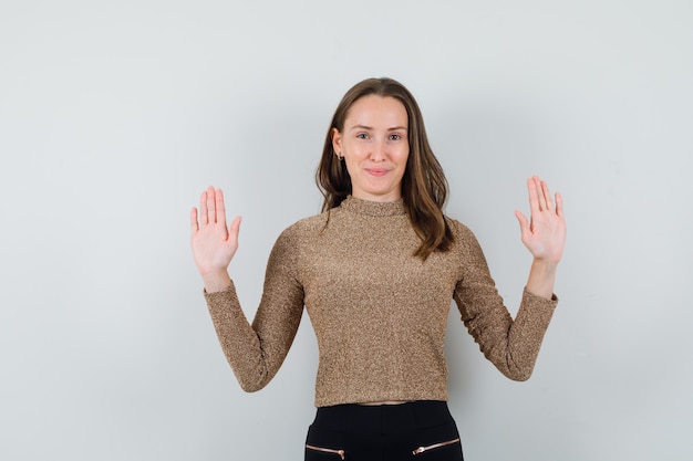 Jeune femme levant les mains tout en montrant ses paumes en chemisier doré et à la recherche de calme. vue de face.